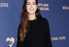 Smiling woman with long wavy hair in a black top posing against a blue Michael Kors backdrop with a heart logo on the left.