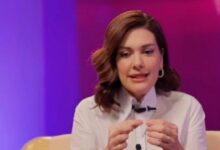 Woman in a white blouse sits on a purple-lit stage, speaking and gesturing with her hands.