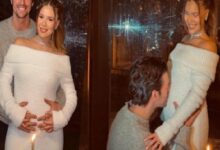 Two women in white off-the-shoulder outfits with two men nearby; one man stands behind the left woman and another kneels to touch the right woman's belly, candlelight in the foreground.