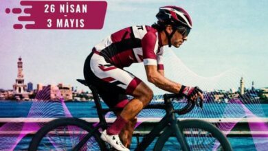 Road cyclist in pink/white kit riding along a waterfront with an event banner reading '26 Nisan 3 Mayıs' in the top left.