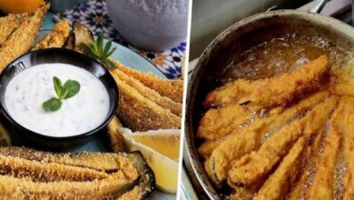 Crispy breaded zucchini sticks with a creamy herb dip in a black bowl and lemon wedges on a decorative plate.