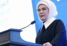 Woman wearing a white hijab and glasses speaks at a black podium with a microphone at a conference booth/stage, blue backdrop behind her.