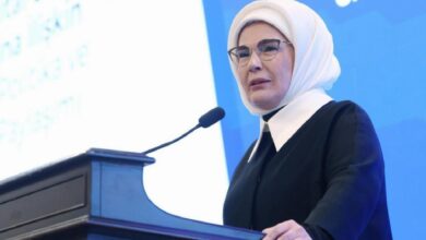 Woman wearing a white hijab and glasses speaks at a black podium with a microphone at a conference booth/stage, blue backdrop behind her.