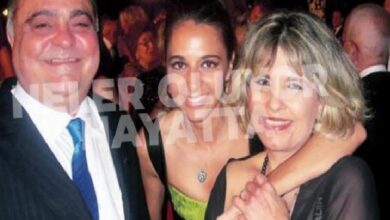 Three adults in formal attire posing together at a crowded social event, with a man in a suit and blue tie on the left and two women embracing in the center and right.