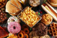 Assorted fast foods and desserts on a table: burgers, fries, donuts, pizza slice, waffles, hot dogs, chocolate bar and more jam-packed together.