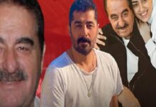 Group of four men posing for a photo: a man in a white T-shirt center, a smiling man with a mustache to the left, and two others leaning in from the right against a red background.