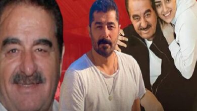 Group of four men posing for a photo: a man in a white T-shirt center, a smiling man with a mustache to the left, and two others leaning in from the right against a red background.