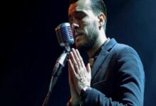 Male singer with hands pressed together in prayer at a microphone on stage.