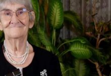 Elderly woman with short gray hair and glasses, wearing a layered pearl necklace and brooch, smiling with green houseplants in the background.