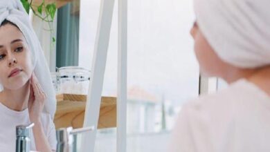 Woman with towel on head looking at her reflection in a bathroom mirror, touching her jawline for skincare