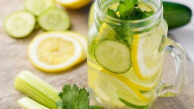 Glass mason jar of lemonade with cucumber, lemon slices and mint leaves, on a wooden table with celery in the foreground
