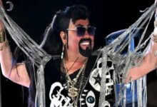 Stage performer wearing sunglasses and a black wig, adorned with multiple gold chains and silver garlands, smiling as they perform.