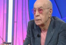Older man with pink-tinted glasses and a pink shirt sits at a TV studio desk wearing a gray blazer and a lapel mic.