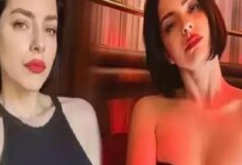 Two women pose for a selfie in a dimly lit setting with red lighting, wearing dark outfits and bold red lipstick.