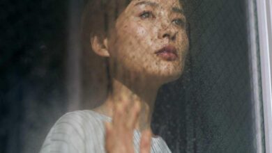 Woman gazing upward through a rain-soaked window, hand pressed against the glass.
