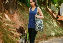 Woman in a blue tank top walks a brown-and-white dog on a lime-green leash along a dirt path, carrying a patterned tote bag over her shoulder.