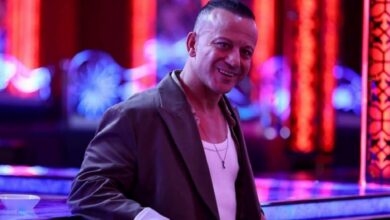 Smiling man in a brown blazer and white tank top leaning on a neon-lit bar in a nightclub setting.