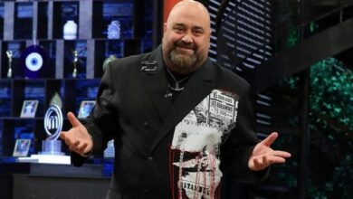 Smiling man in a black double-breasted blazer with a newspaper-print panel on the left, gesturing on a television studio set.