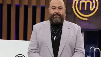 Bearded, bald man in a light gray blazer and black shirt on a TV studio set with a MasterChef logo in the background.