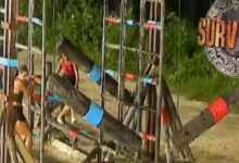Survivor challenge on a sandy beach with tall wooden frames wrapped in blue and red tape and contestants in swimsuits, Survivor logo in the top right.
