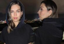Two women in black hoodies on a rooftop balcony at night; one faces the camera while the other is in profile with city lights in the background.