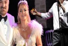 Bride in a white beaded gown and tiara stands beside a man in a white tuxedo on a stage, with a singer performing in the background.