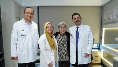 Four people in a modern exam room: two doctors in white coats with ID badges flank a smiling patient and a woman in a leopard-print top, all posing for a photo.