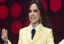 Woman on a stage wearing a pale yellow suit and black tie, holding a microphone and smiling.